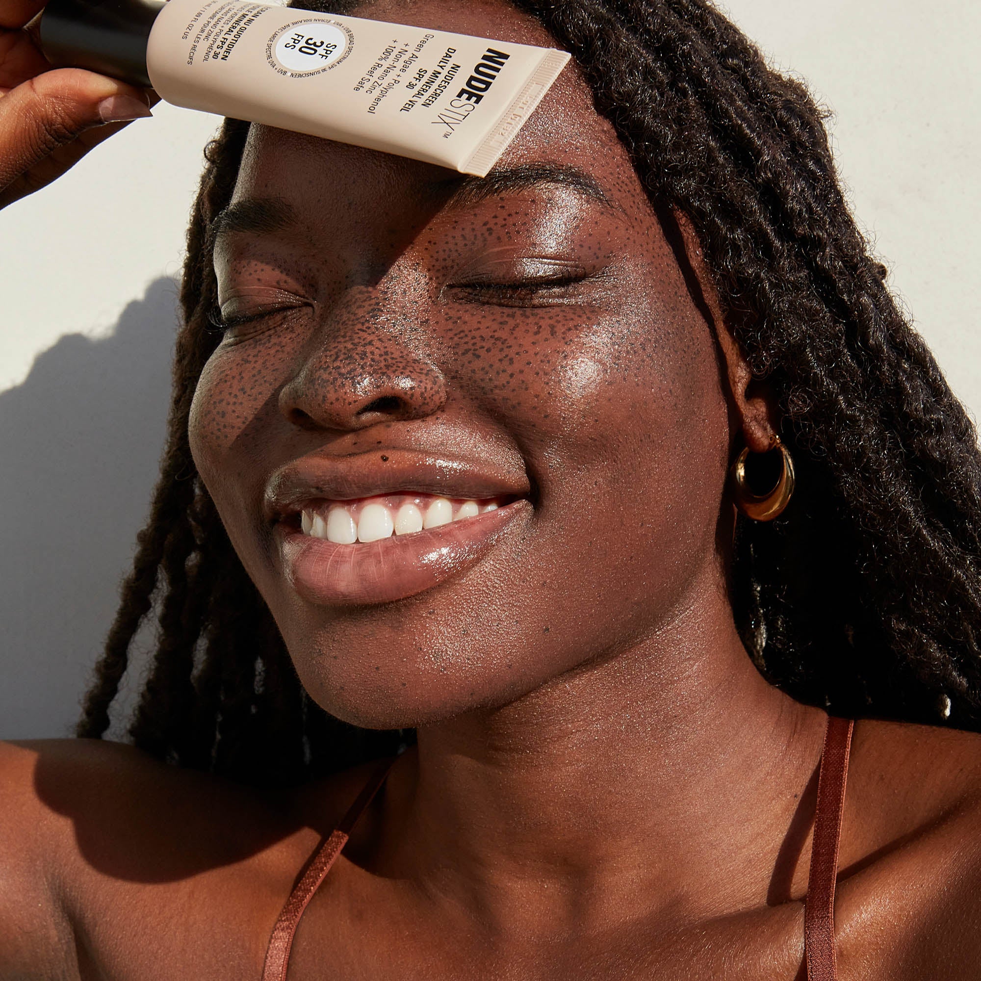 Dark skinned freckled young woman smiling while holding a tube of Nudescreen Daily Mineral Veil